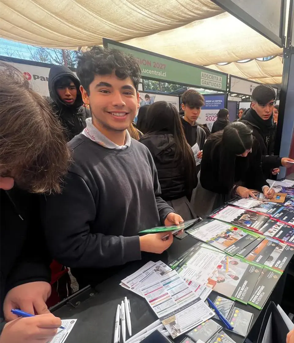 Estudiante sonríe mientras participa en feria educativa junto a compañeros y expositores. Estudiante sonríe mientras participa en feria educativa junto a compañeros y expositores.